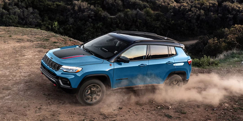A blue Jeep Compass driving off-road, kicking up dust.